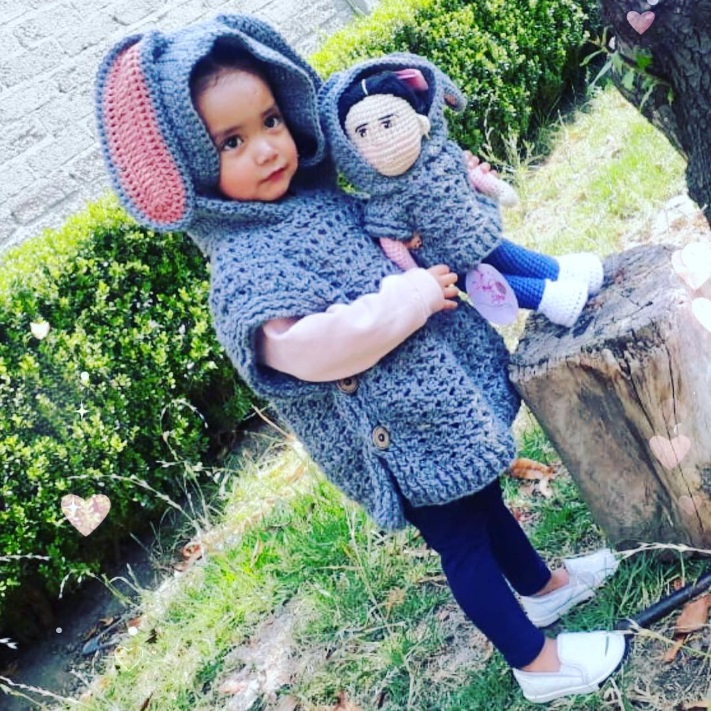 A kid wearing a crochet rabbit jacket, holding a replica of herself wearing the same jacket